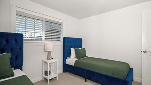 A bedroom with two beds, a window with blinds, a bedside table with a lamp, and a door. - Updated Spacious Home & Hot Tub (Spokane Valley)