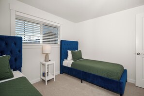 A bedroom with two beds, a window with blinds, a bedside table with a lamp, and a door.