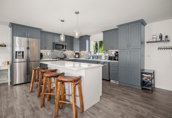 A modern kitchen with grey cabinets, a stainless steel refrigerator, a large island with bar stools, and a window providing natural light.