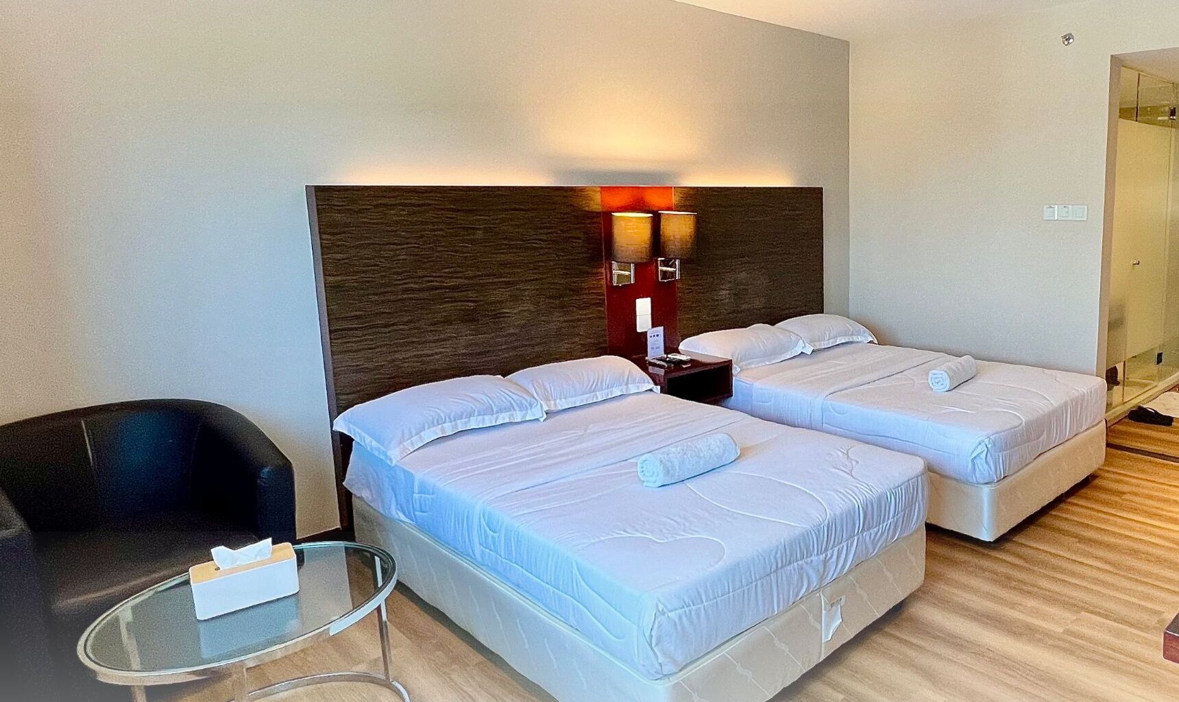 A hotel room with two beds, a small table, a chair, and a wall-mounted lamp.