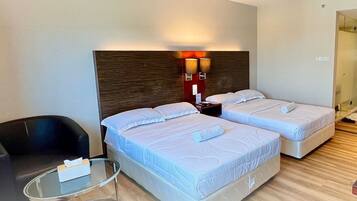 A hotel room with two beds, a small table, a chair, and a wall-mounted lamp.