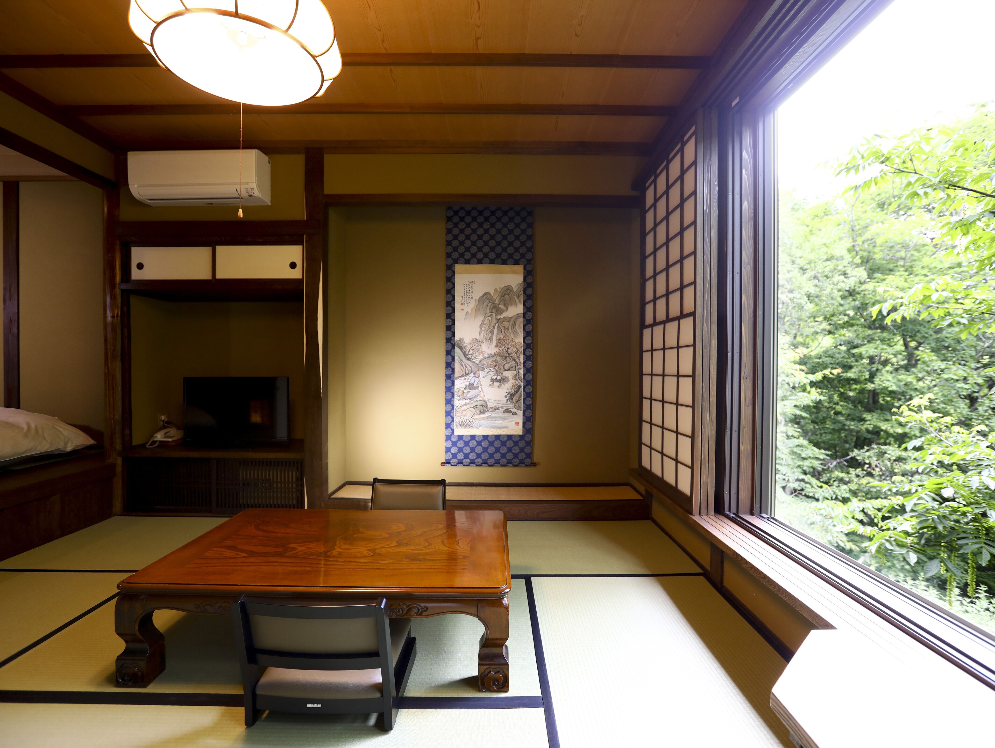 Una habitación japonesa tradicional con suelo de tatami, una mesa de madera baja, un televisor y un ventanal con vistas a la vegetación.