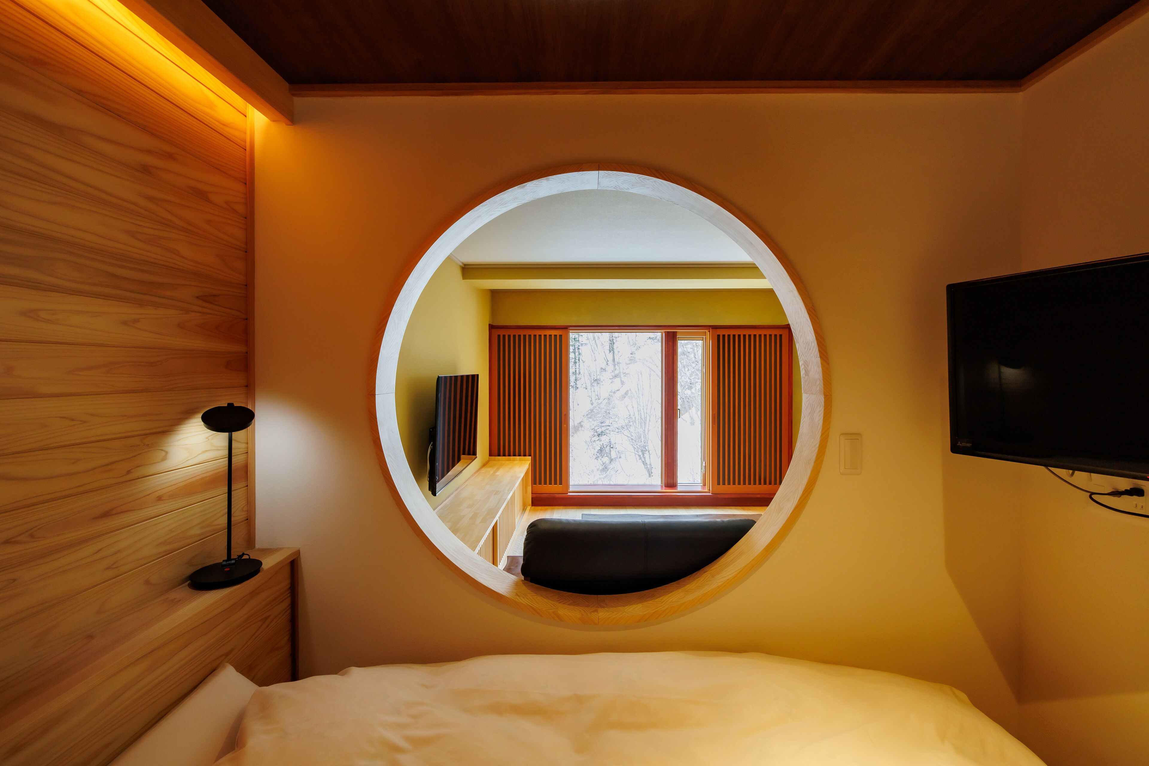 A bedroom with a circular window, a bed, a bedside table, a lamp, and a TV mounted on the wall.