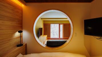 A bedroom with a circular window, a bed, a bedside table, a lamp, and a TV mounted on the wall.