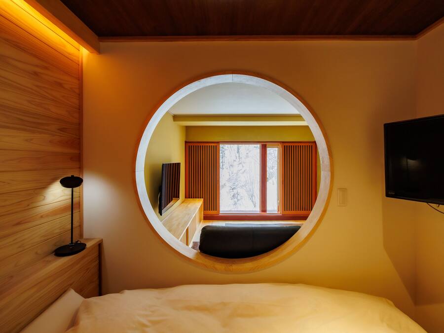 A bedroom with a circular window, a bed, a bedside table, a lamp, and a TV mounted on the wall.
