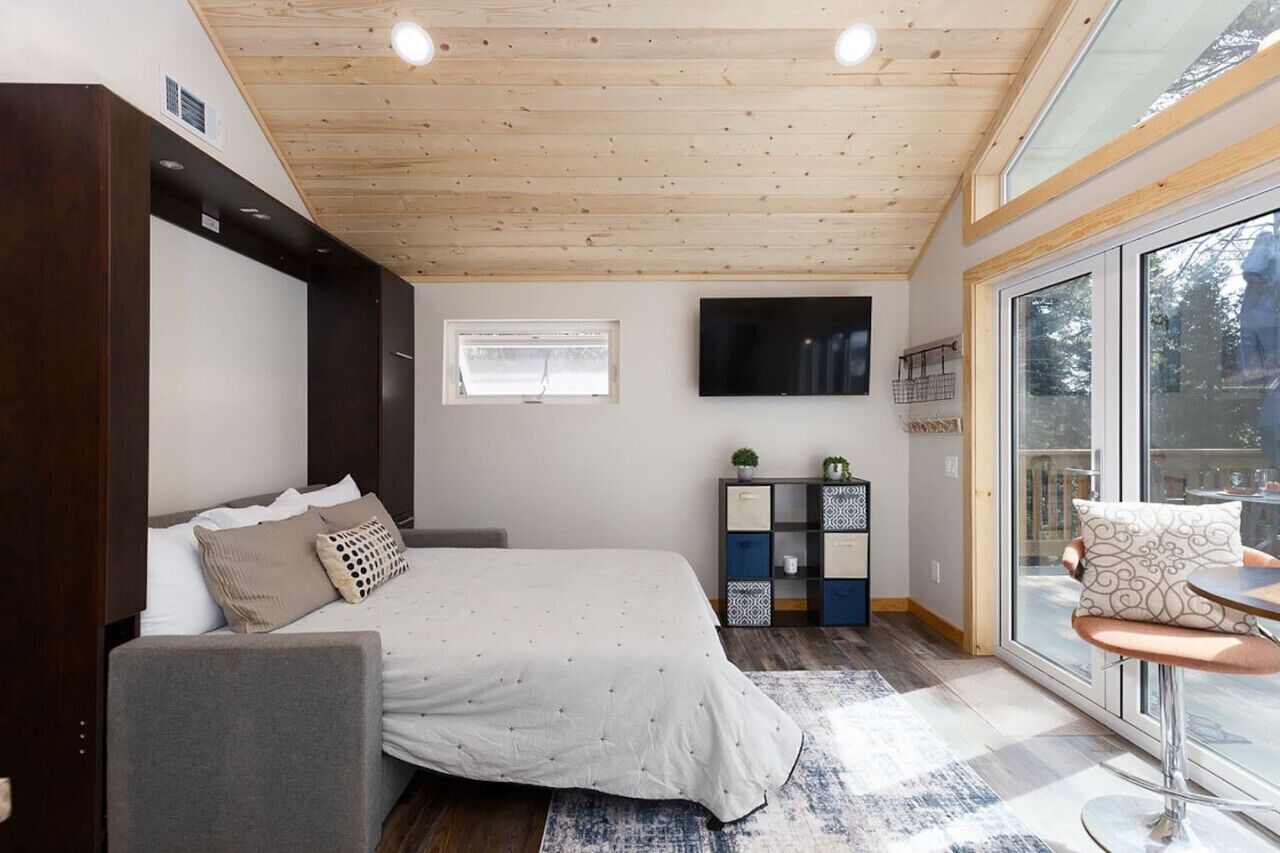 A bedroom with a bed, a sofa, a TV, a window, and a sliding glass door to a balcony.