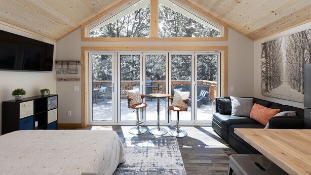 A spacious room with a high ceiling, a large sliding glass door leading to an outdoor deck, a bed, a sofa, and a dining table. - Bugsy’s Escape House|Private|Nature (Lake Arrowhead)