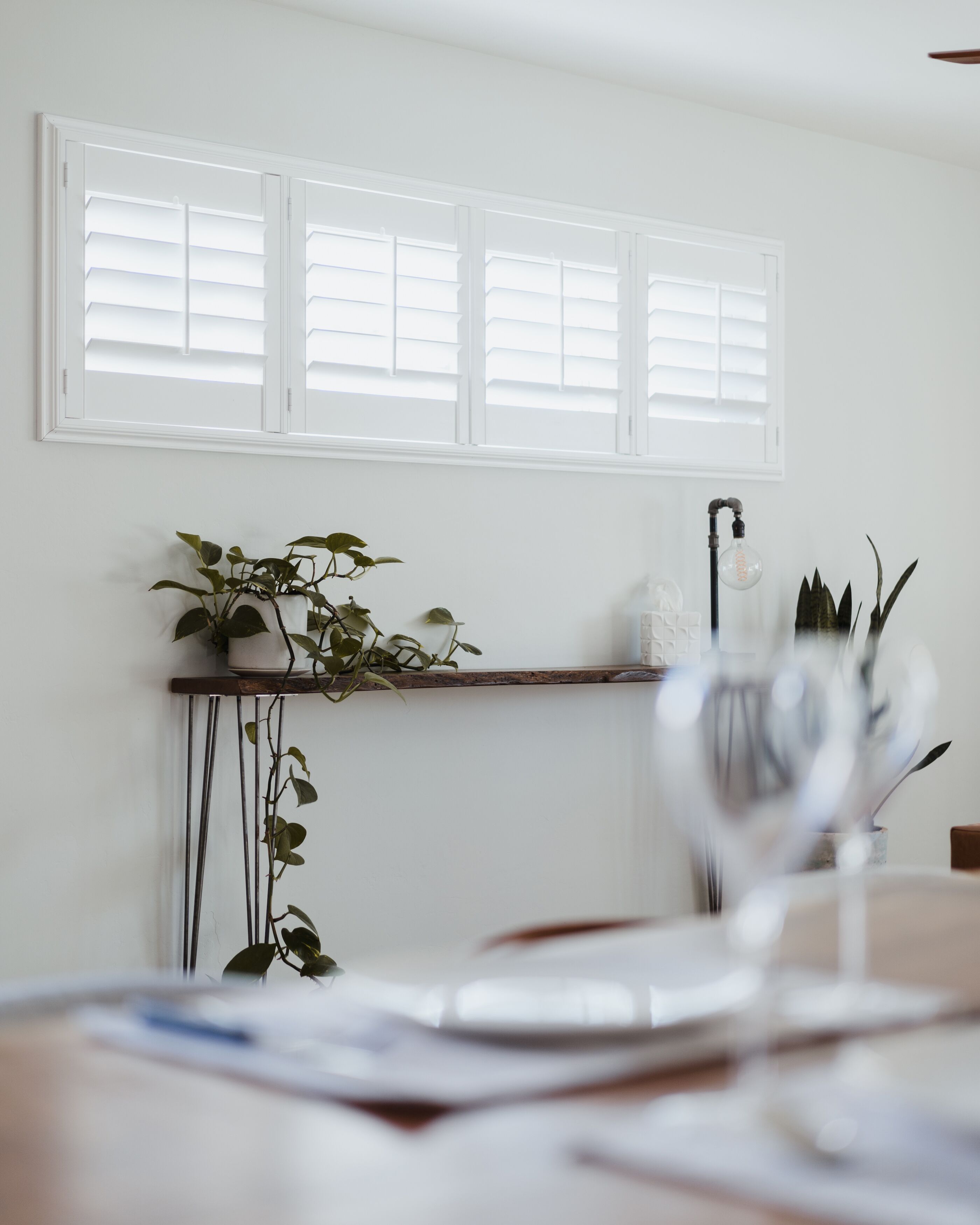 Una habitación con pared blanca, una ventana con persianas, una estantería con plantas y una mesa con papeles y un vaso.