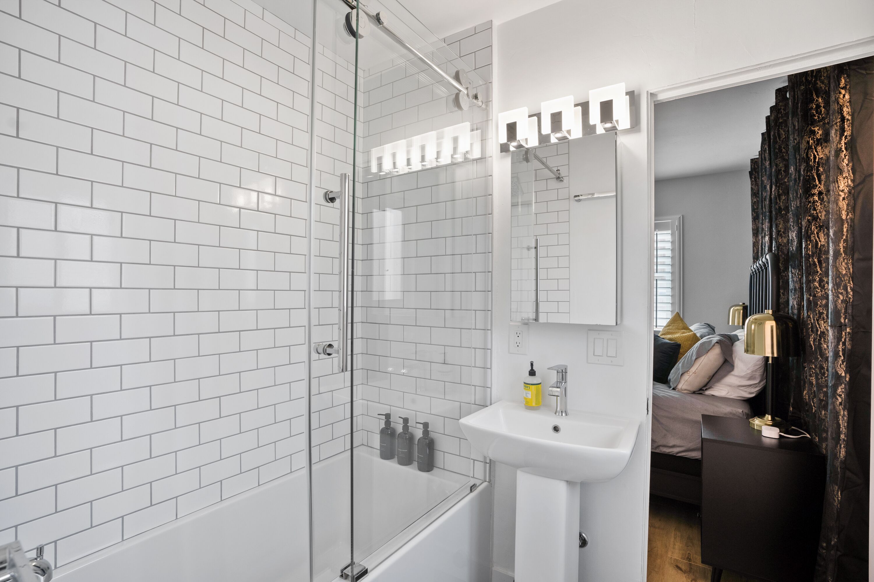 Een moderne badkamer met witte metrotegels, een glazen douchecabine, een witte wastafel en een nachtkastje met een lamp.