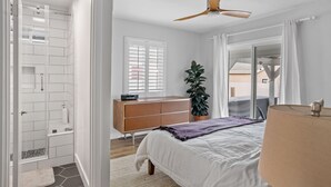 Een slaapkamer met een bed, een dressoir, een plant, een raam met jaloezieën en een plafondventilator.