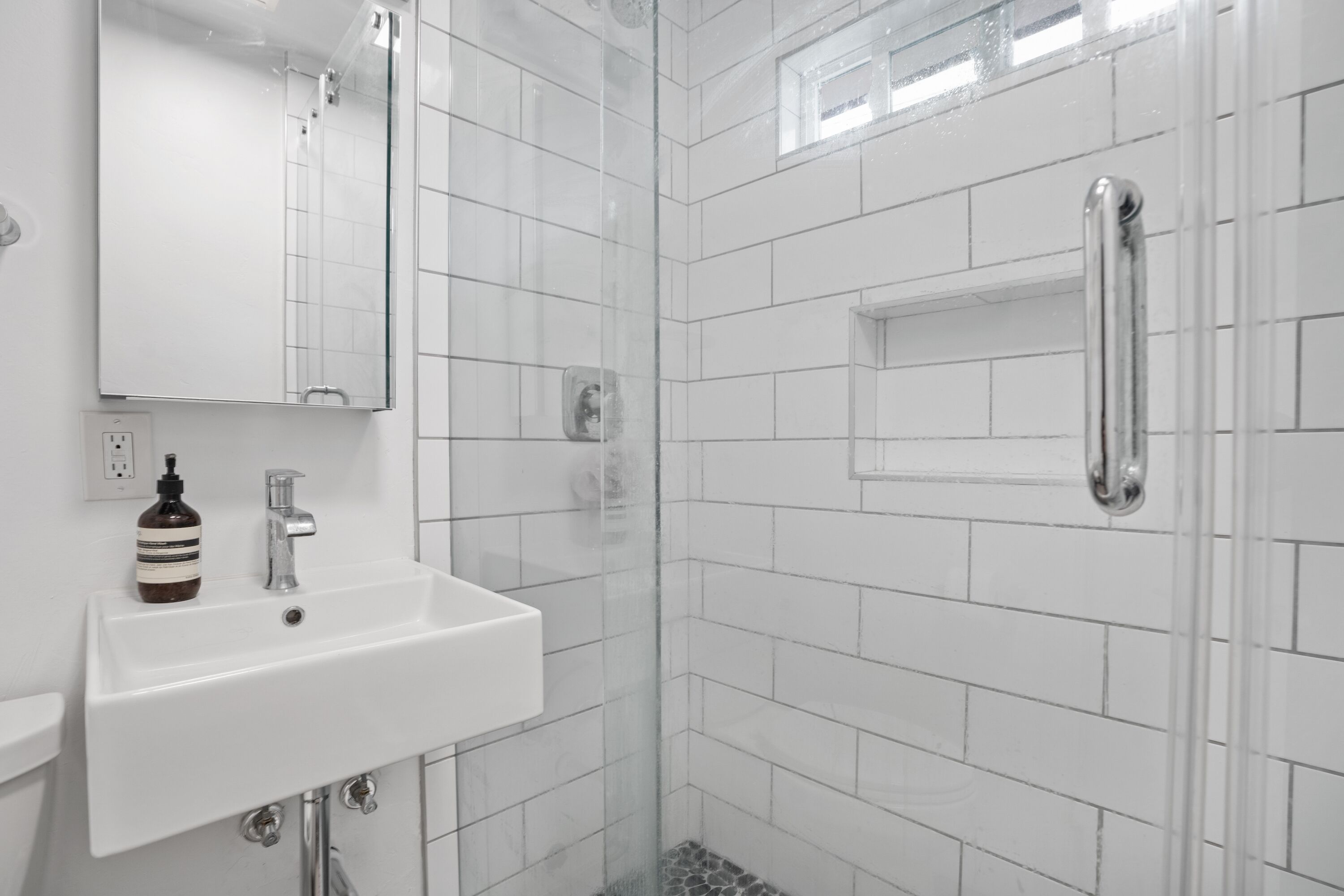 A modern bathroom with white tiles, a glass-enclosed shower, a white sink with a soap dispenser, and a mirror above the sink.