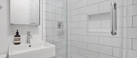 A modern bathroom with white tiles, a glass-enclosed shower, a white sink with a soap dispenser, and a mirror above the sink.