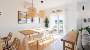 Un salon lumineux avec une table à manger en bois, des chaises blanches, un canapé et des fauteuils en osier. On y trouve deux lampes en osier suspendues, un grand miroir et une plante en pot.