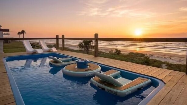 Een zwembad met ligbedden en een houten terras met uitzicht op de oceaan bij zonsondergang.