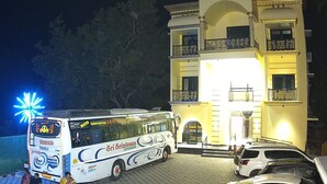 A white bus with "Sri Srinivasa Travels" branding parked in front of a building with multiple balconies and a well-lit entrance.