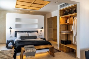 A modern bedroom with a bed, bedside tables, a bench, a wardrobe, and a built-in heater. - Arotron Santorini (Santorini)