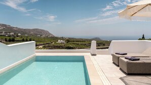 A pool with a view of a coastal landscape, a white building, and a lounge area with a sofa and umbrella. - Arotron Santorini (Santorini)