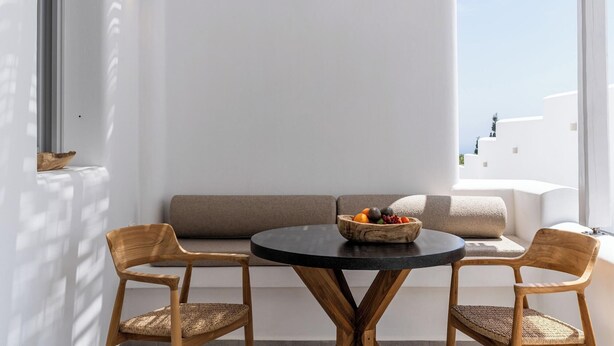 A balcony with a round table, two wicker chairs, a wooden bowl with fruits, and a white wall with a window. - Arotron Santorini (Santorini)