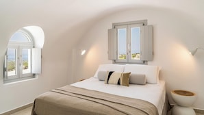 A bedroom with a bed, a window with a view, a bedside table, and a lamp. - Arotron Santorini (Santorini)