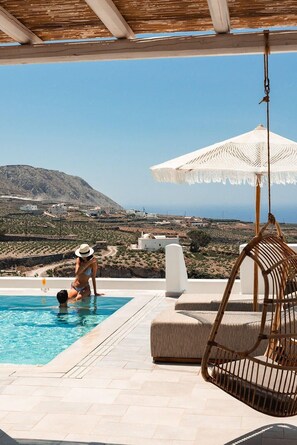 A pool area with a view of a mountainous landscape, a hanging chair, and a white umbrella. - Arotron Santorini (Santorini)