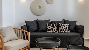 A modern living room with a black sofa, wooden chair, and two woven side tables. - Arotron Santorini (Santorini)