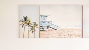 A bedroom with a white headboard, a bed with blue and green pillows, and a wall-mounted artwork depicting a beach scene with palm trees and a lifeguard tower.
