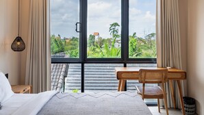 Une chambre à coucher avec un lit, un bureau avec des chaises, et une vue sur la verdure et les bâtiments, visible à travers de grandes fenêtres.