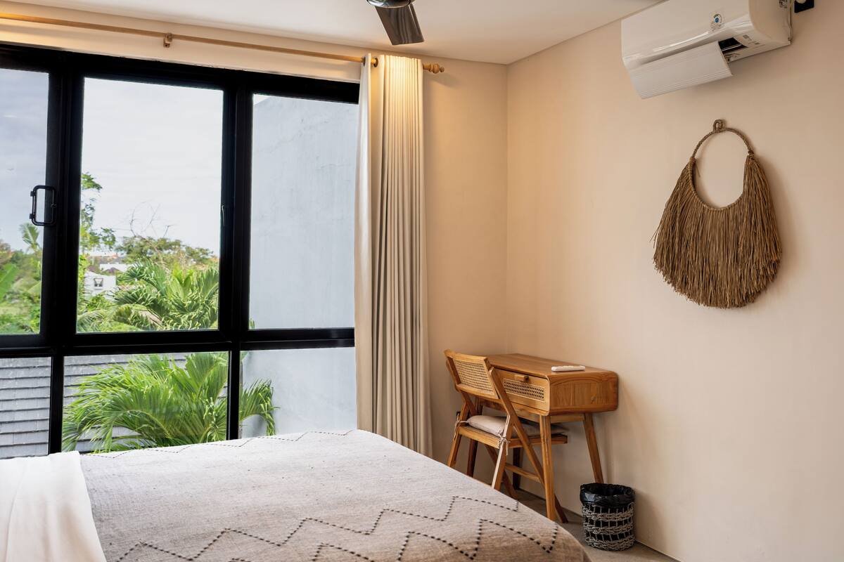 Quarto de hotel com cama, escrivaninha, cadeira, ar condicionado fixado na parede e vista para o verde lá fora.