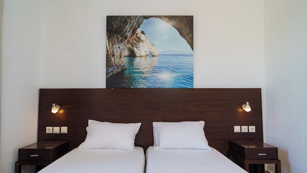A hotel room with two beds, a wooden headboard, bedside lamps, and a painting of a coastal scene. - Mayo Rooms (Zakynthos)