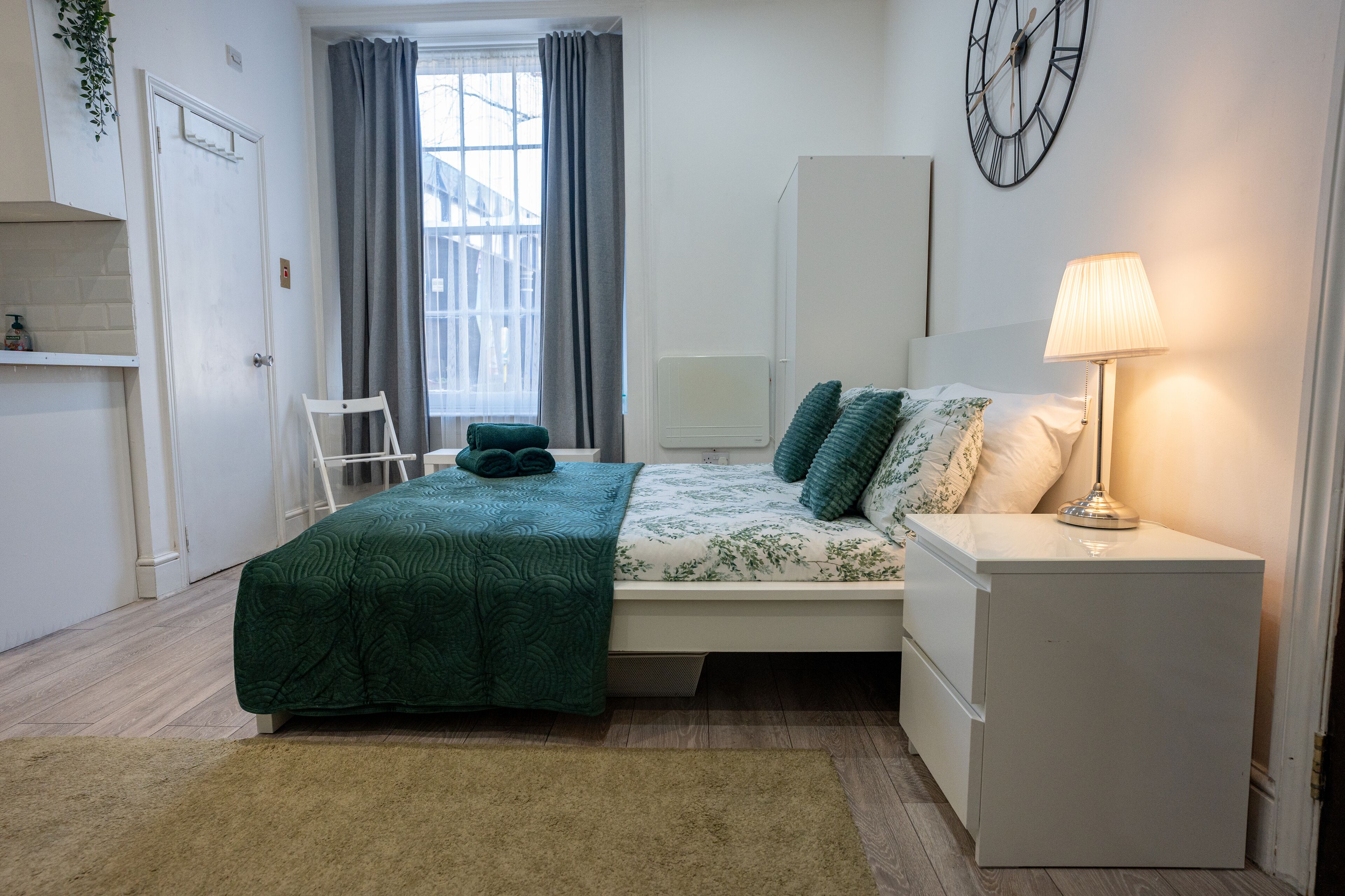 A bedroom with a bed, bedside table, lamp, and a clock on the wall.