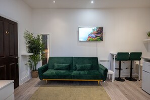 Un salon moderne avec un canapé vert, un téléviseur à écran plat, une plante en pot et un petit coin repas avec deux chaises.