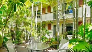 Un patio con una mesa y sillas de vidrio, rodeado de exuberante vegetación tropical y un edificio de varios pisos con balcones.