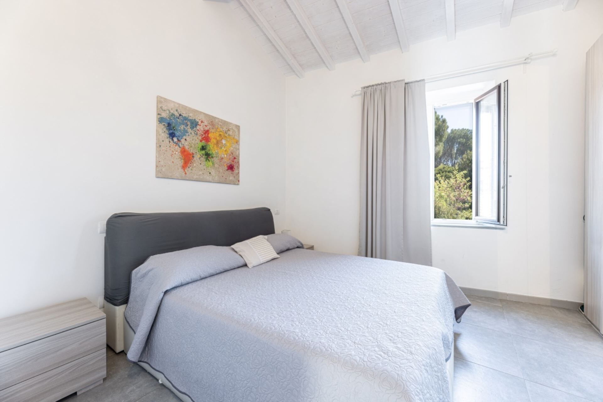 A bedroom with a bed, a nightstand, a window with curtains, and a painting on the wall.