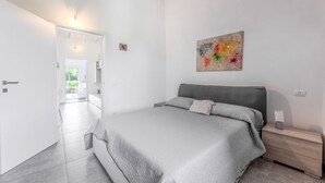 A bedroom with a bed, a bedside table, a nightstand, a painting on the wall, and a door leading to another room. - Casetta Ginevra Country & Modern (Portoferraio)