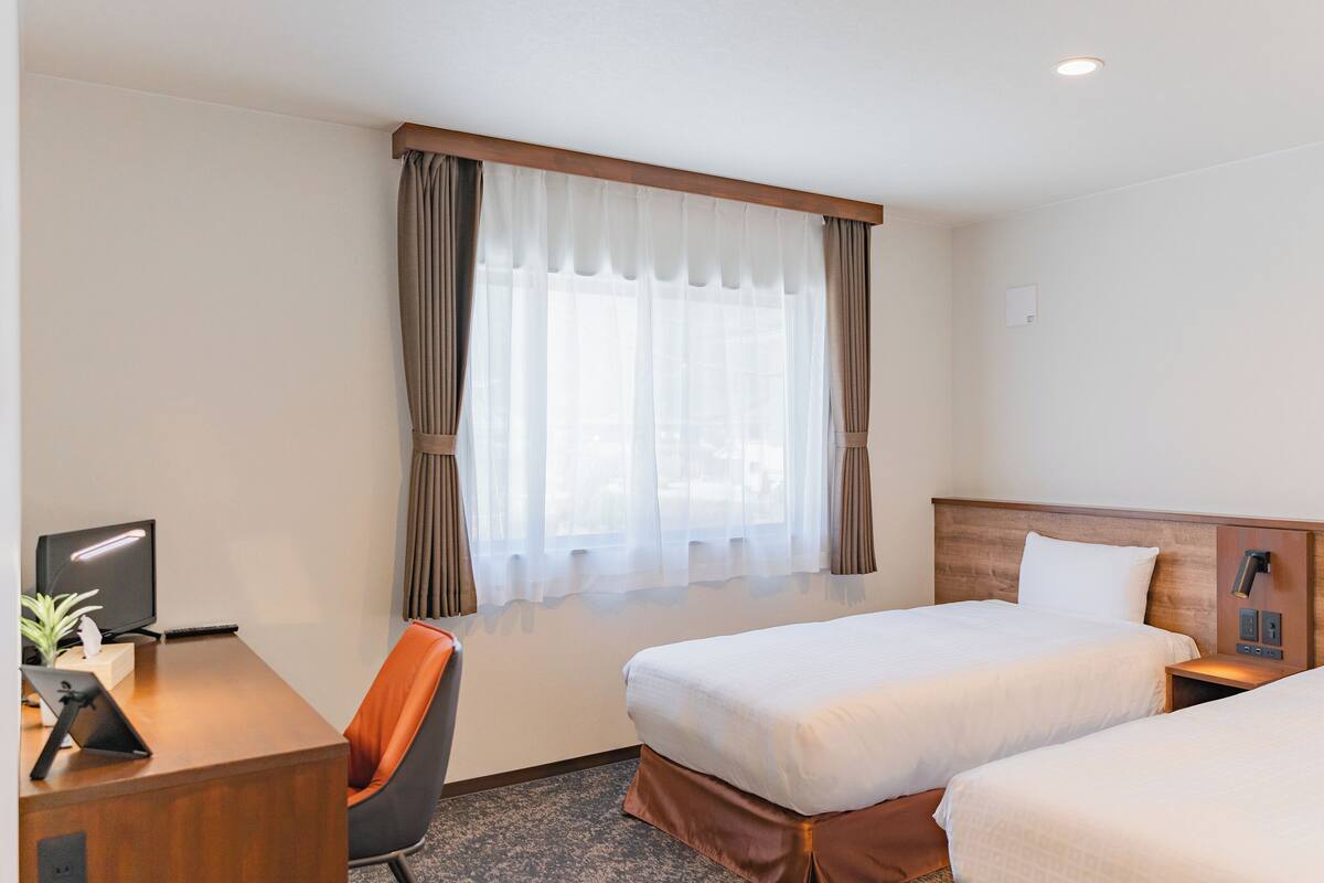 A hotel room with two beds, a desk with a chair, a computer, and a window with curtains.
