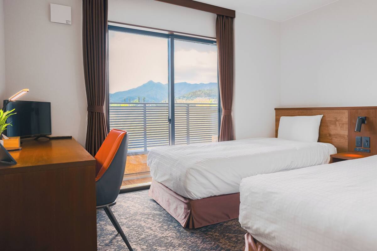 A hotel room with two beds, a desk with a computer, a chair, and a view of mountains through the window.