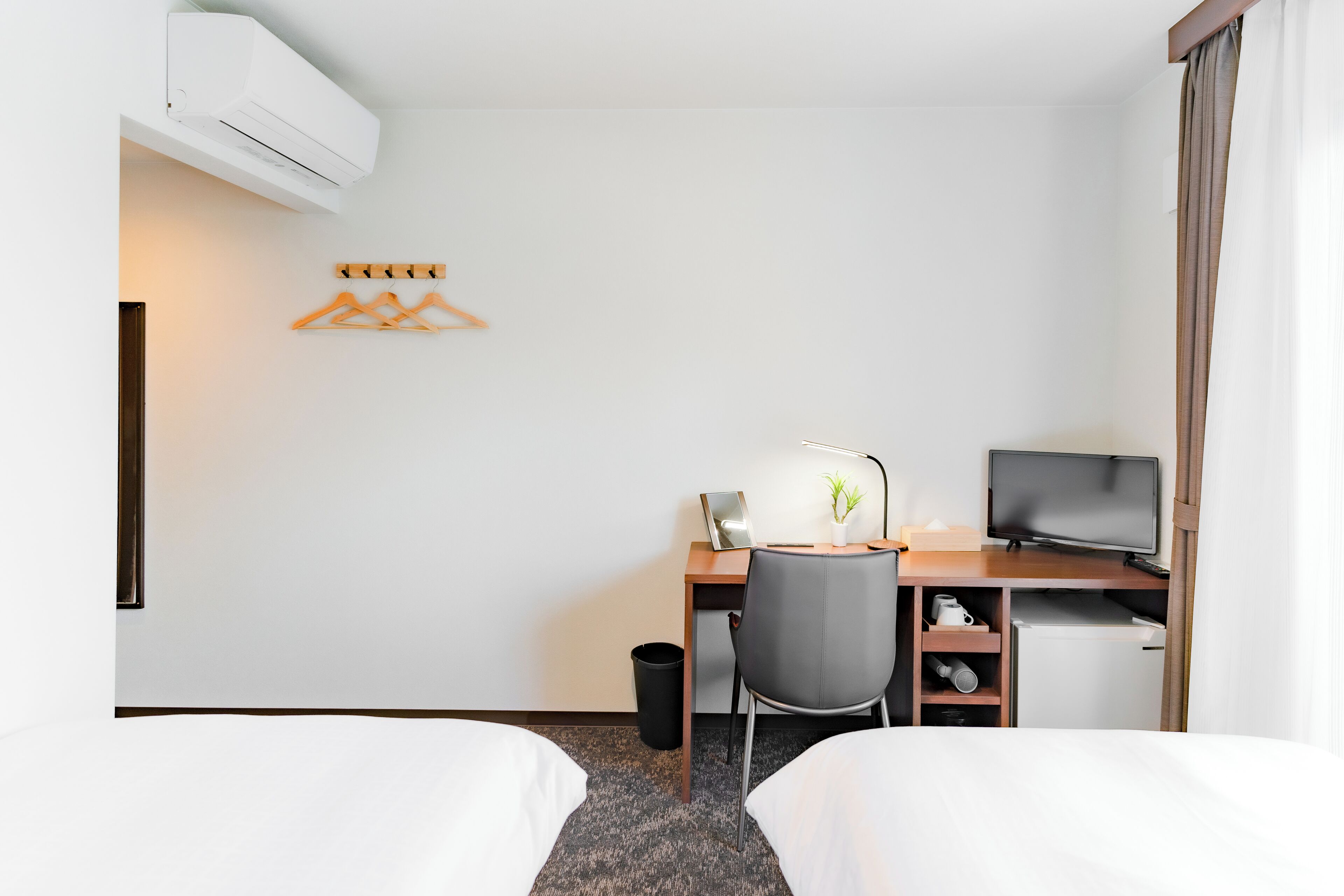 A hotel room with two beds, a desk with a computer and a TV, a small fridge, a desk lamp, and a plant.