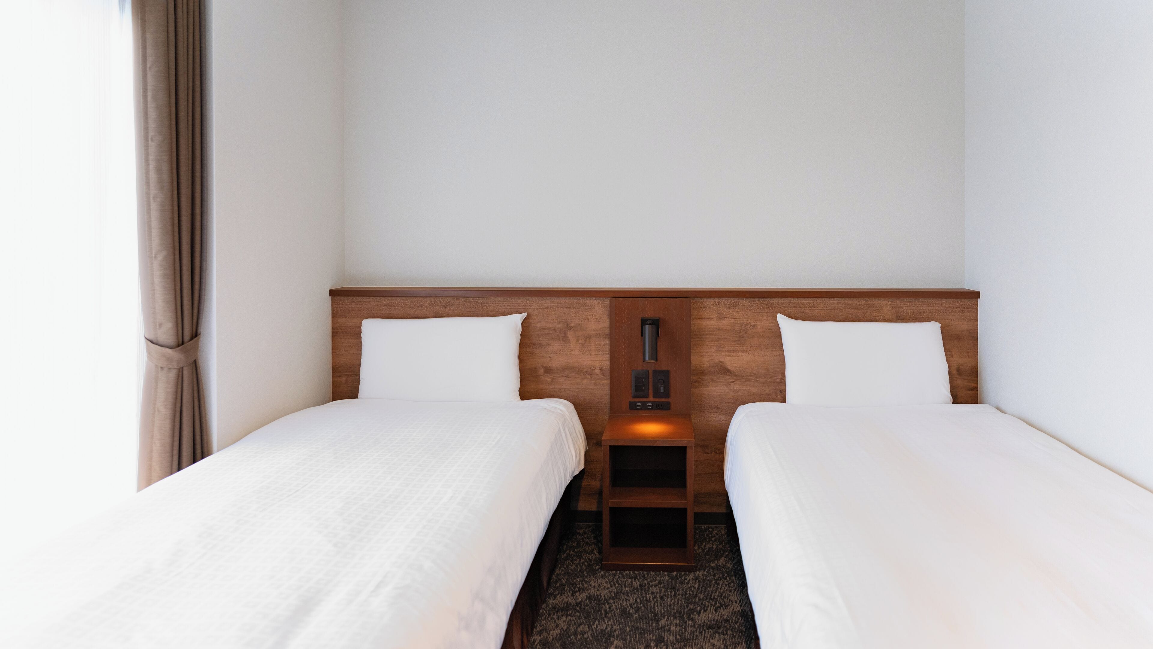 A hotel room with two single beds, a wooden headboard, a bedside table with a lamp, and a window with curtains.