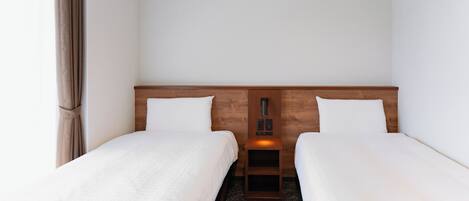 A hotel room with two single beds, a wooden headboard, a bedside table with a lamp, and a window with curtains.