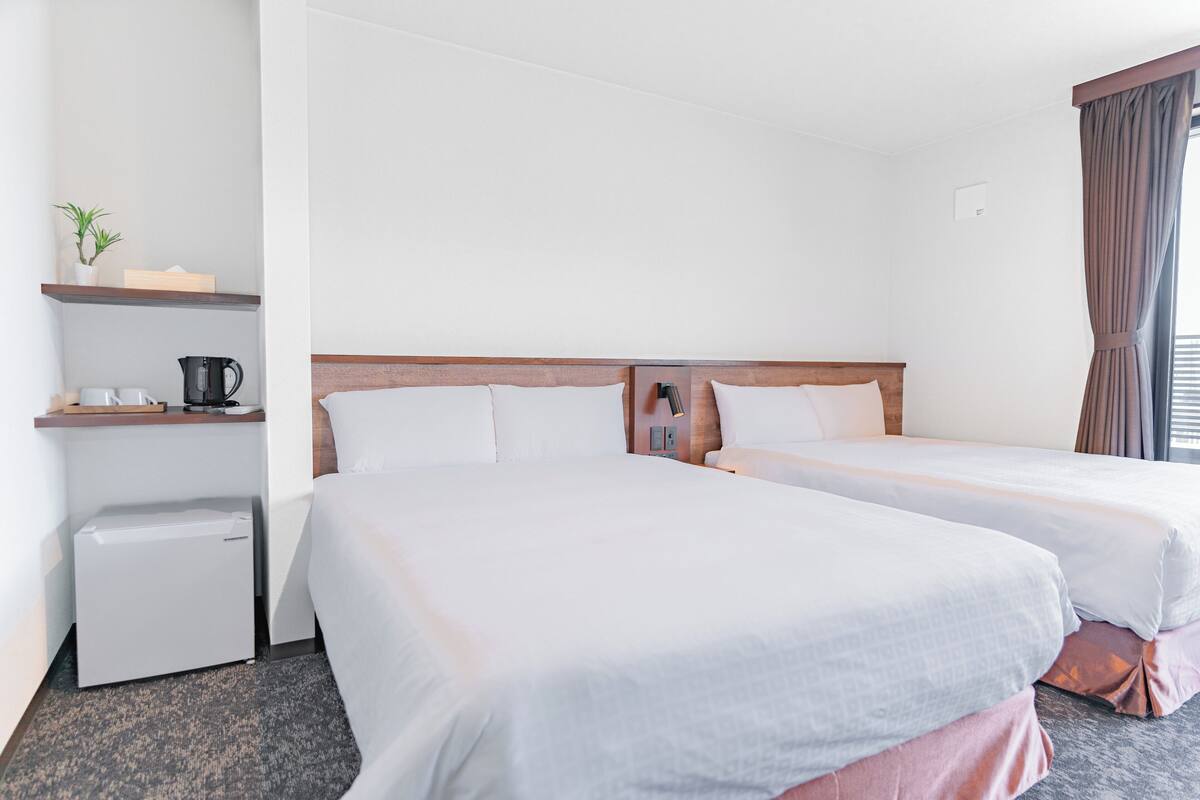 A hotel room with two beds, a small desk with a kettle, a plant, and a window with curtains.