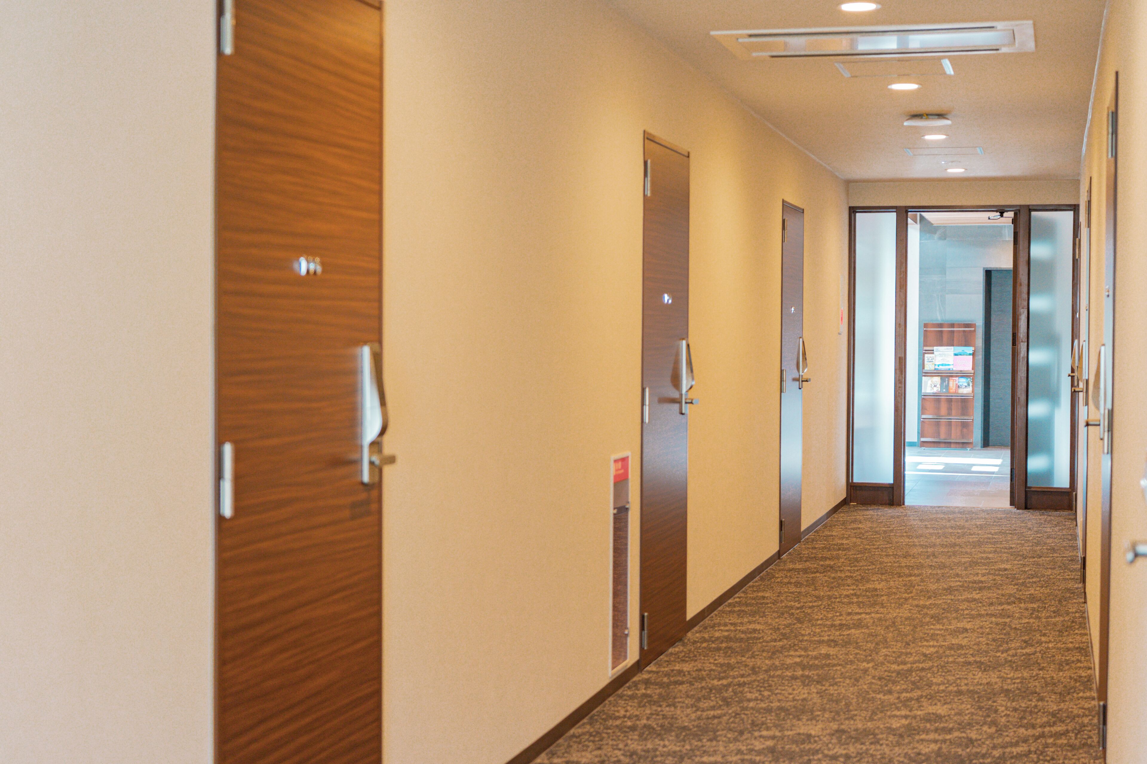 A hotel corridor with wooden doors, a carpeted floor, and a view into a room with a desk and chair.