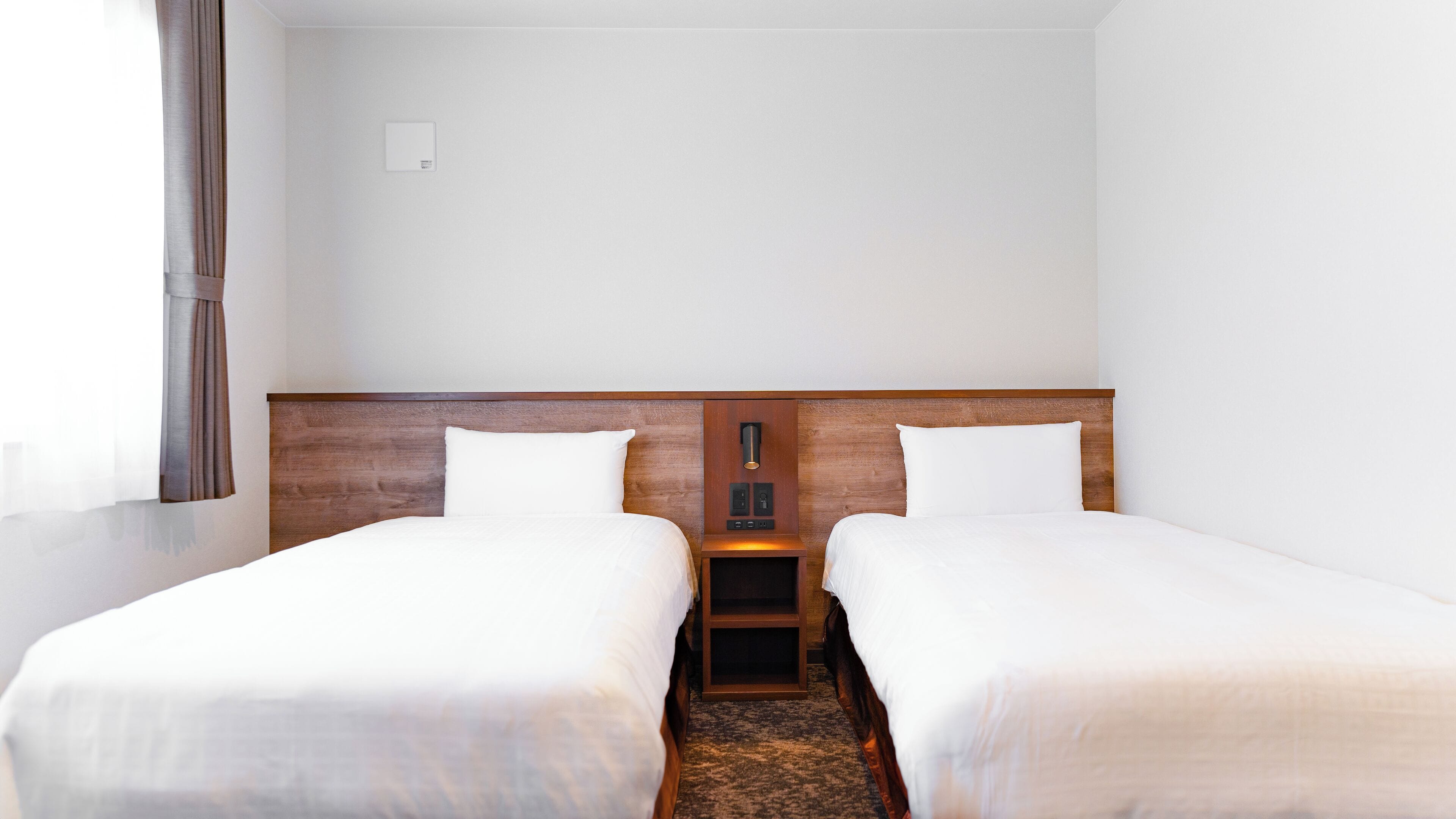 A hotel room with two single beds, a wooden headboard, a nightstand with a lamp, and a plain white wall.