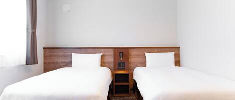 A hotel room with two single beds, a wooden headboard, a nightstand with a lamp, and a plain white wall.