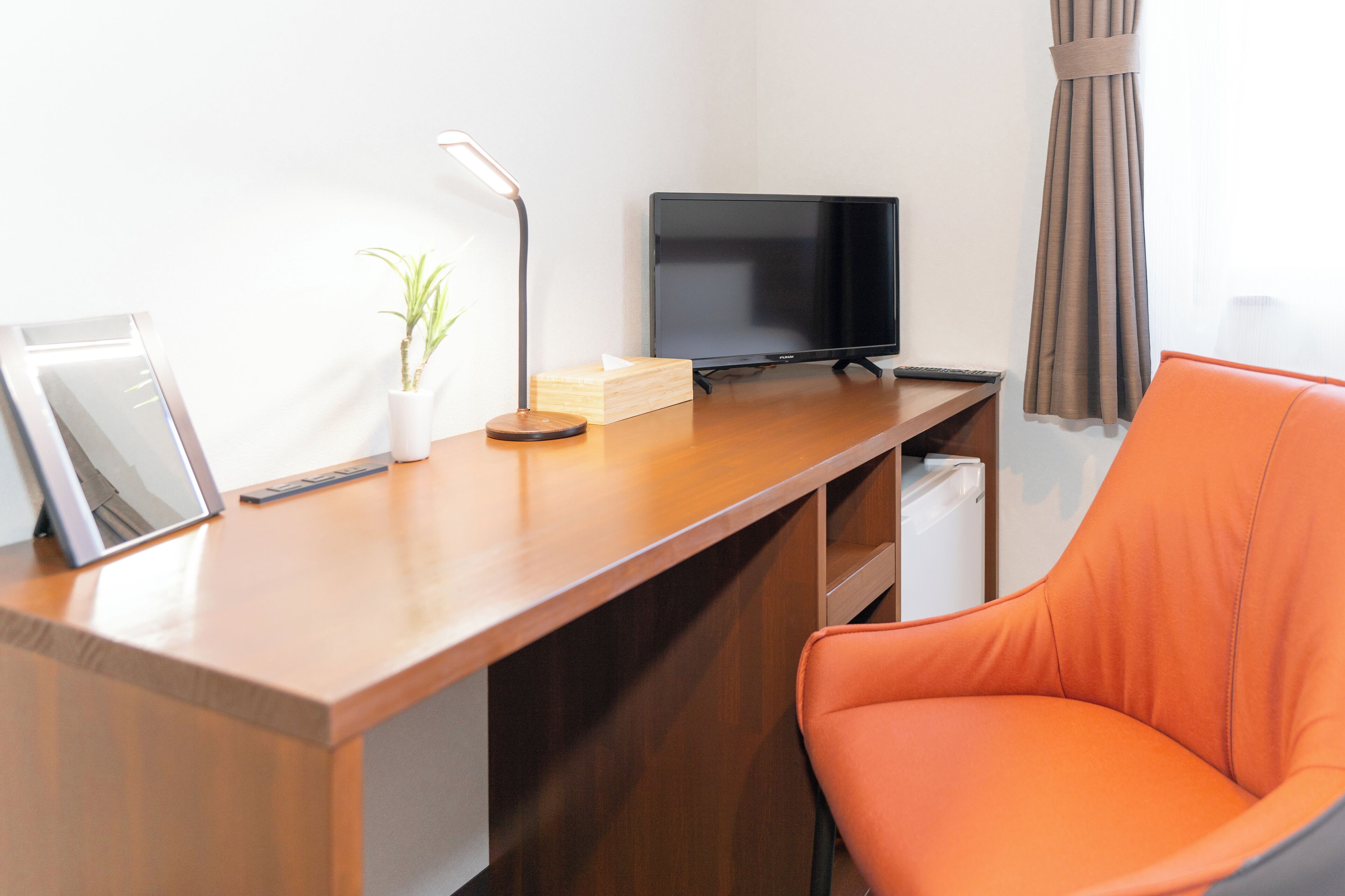 Une chambre d’hôtel avec un bureau en bois, un téléviseur noir, une plante en pot, un tableau encadré et une chaise orange.