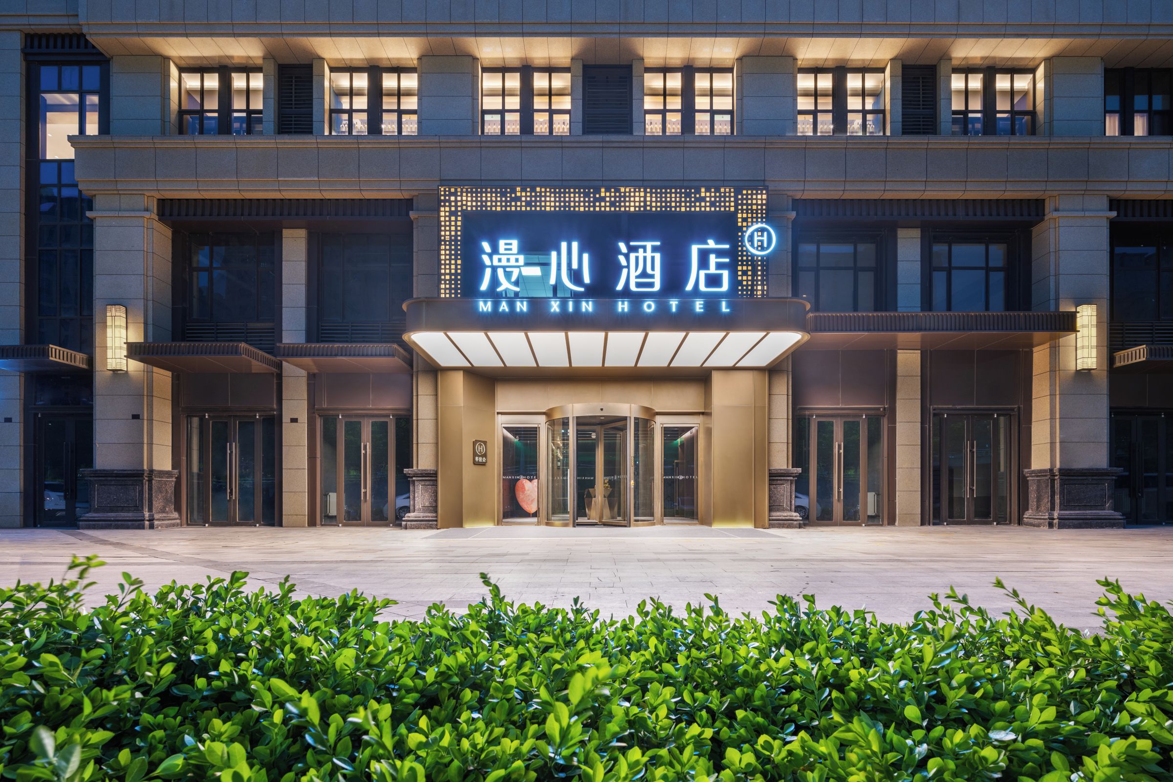 A hotel entrance with a sign in both Chinese and English, a revolving door, and a well-maintained garden area in front.