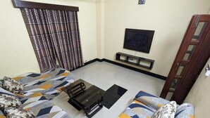 A living room with a sofa set, a coffee table, a flat-screen TV mounted on the wall, and a window with patterned curtains. - Rainagar rajbari (Sylhet)