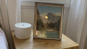Uma mesa de cabeceira com um quadro emoldurado, uma luminária branca e uma mesinha de madeira.