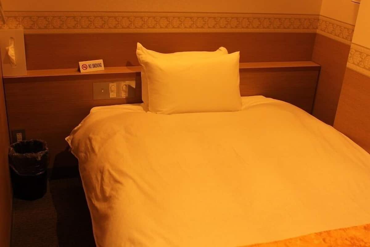A hotel room with a bed, a bedside table, a trash bin, and a sign indicating no smoking.