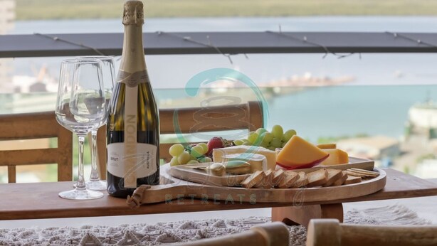 Una botella de vino Brut, una copa, y una tabla de madera con uvas, queso y galletas en un balcón con vista al agua.