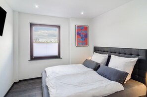 Une chambre à coucher avec un lit, une fenêtre avec des stores, un tableau accroché au mur et un luminaire au plafond.